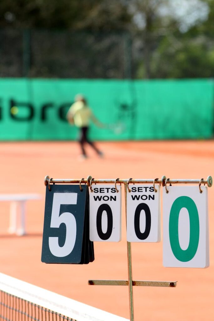 pexels-photo-899318 Close-up of tennis scoreboard with blurred player in action on clay court.